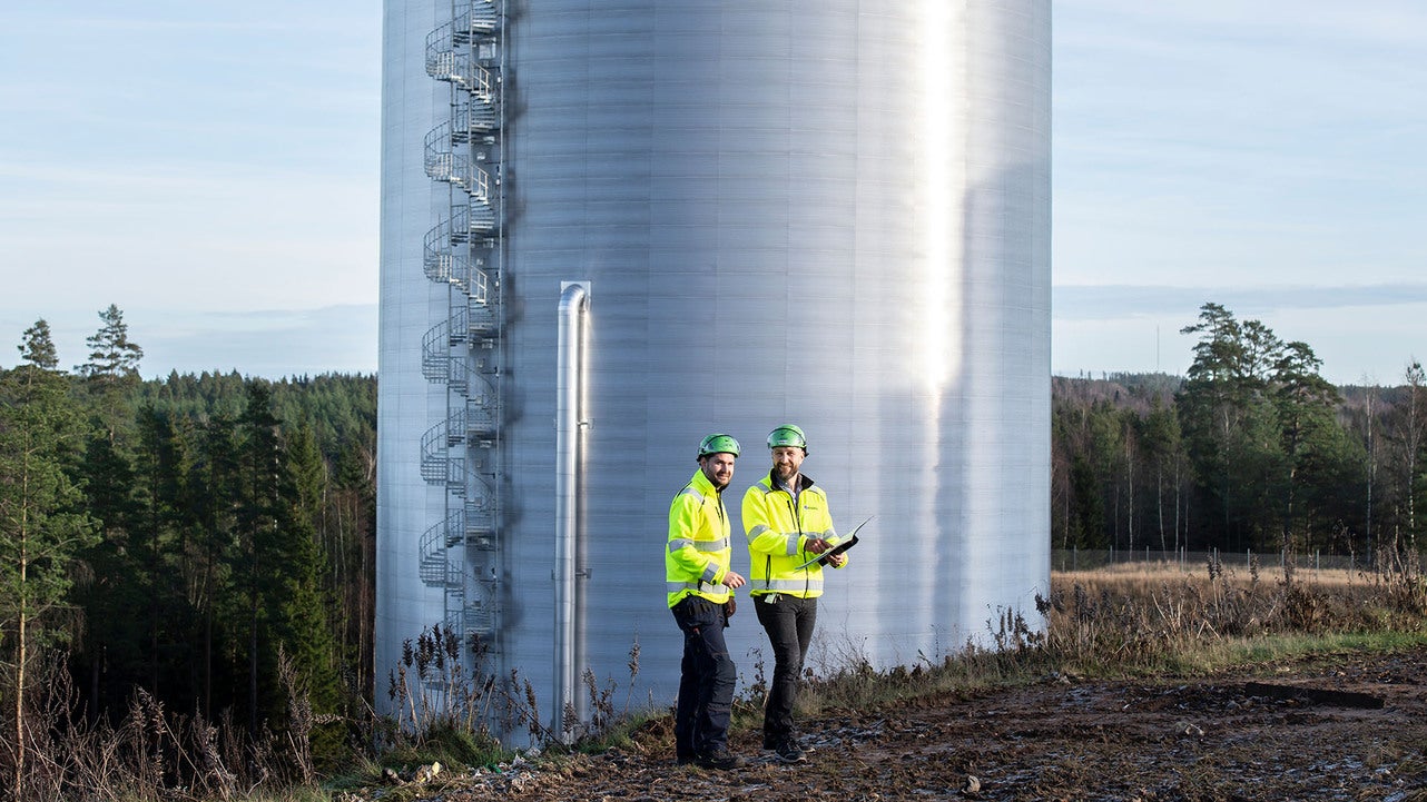 Två tekniker från Jönköping Energi, klädda i gula skyddsjackor och hjälmar, står framför en stor, silverfärgad ackumulatortank utomhus. De håller i arbetsdokument och befinner sig vid kanten av ett skogsområde.