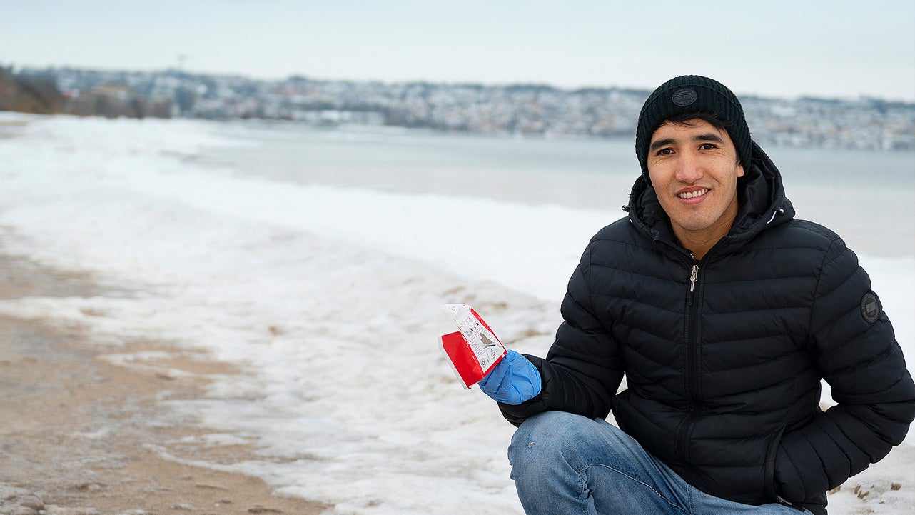 En person i svart vinterjacka och mössa sitter vid en strand med snö, nära sjön Vättern, och håller upp skräp som samlats in i en röd påse. Staden syns i bakgrunden.