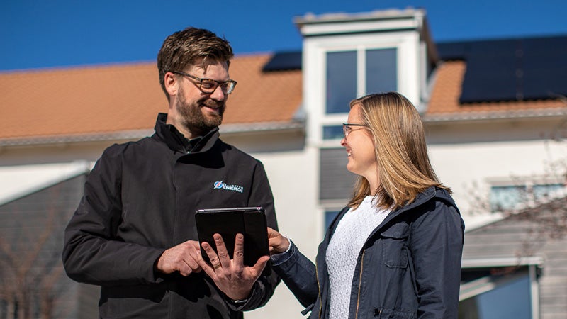 En manlig medarbetare från Jönköping Energi står med en surfplatta och diskuterar med en kvinna framför ett hus med solpaneler på taket. Båda ler medan de tittar på skärmen, och en klarblå himmel syns i bakgrunden.