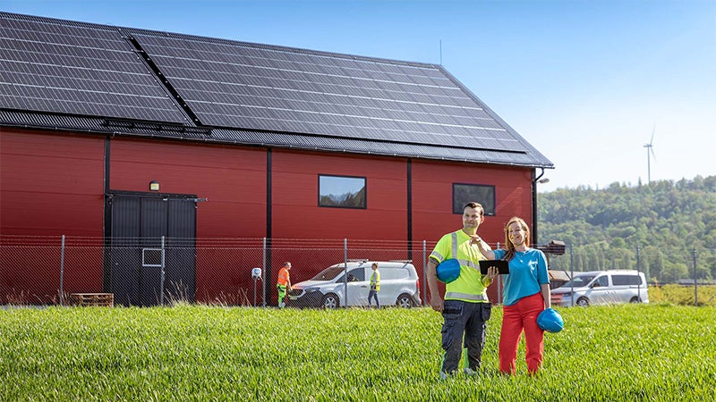 Två personer står framför en röd byggnad med solpaneler på taket, omgivna av gräsfält och fordon, med ett vindkraftverk synligt i bakgrunden.