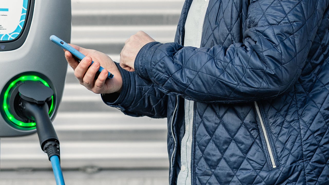 En person i en blå jacka står vid en laddstation för elbilar och håller en smartphone. Laddstationens kabel är ansluten till bilen, och en grön lysring runt anslutningen indikerar att laddning pågår.