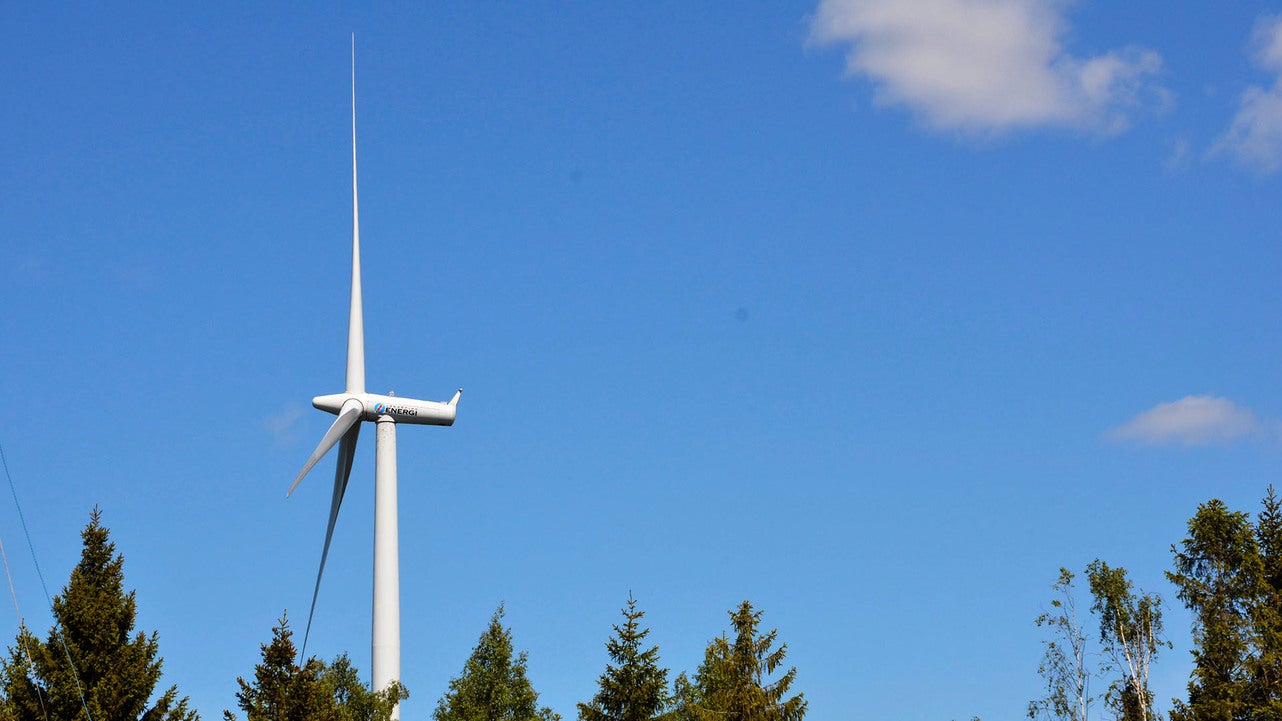 Ett vindkraftverk reser sig över trädtopparna på en klarblå himmel med några få moln. Vindkraftverkets vita rotorblad är i rörelse och kontrasterar mot den blå himlen.