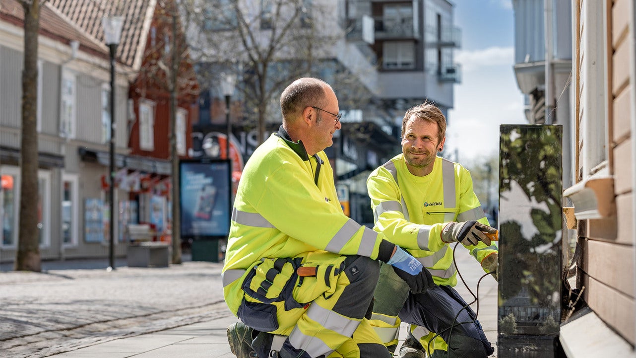 Två tekniker från Jönköping Energi, klädda i gula arbetskläder, arbetar på en elinstallation utomhus i en central del av Jönköping. De sitter vid en byggnad och har verktyg i händerna, omgivna av stadsmiljö med butiker och gångvägar.