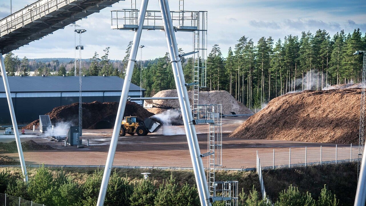 En biobränsleanläggning på Torsvik med stora högar av träflis och en gul hjullastare som arbetar på området. Anläggningen är omgiven av skog och industribyggnader, med transportband och rör som syns i förgrunden.