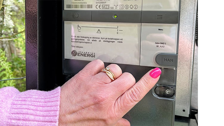 Hand med rosa nagellack trycker på HAN-knappen på en digital elmätare märkt Jönköping Energi, med display och informationstext synlig.