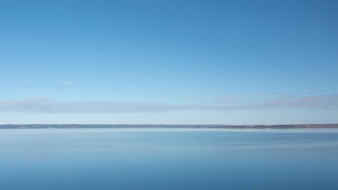 En vidsträckt vy över den lugna sjön Vättern, där himlen och vattnet smälter samman i blå nyanser. Horisonten är tydlig och sträcker sig över hela bilden, vilket skapar en känsla av ro och öppenhet.