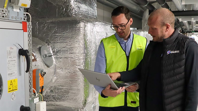 två medarbetare från jönköping energi står i en industriell miljö och diskuterar samtidigt som de kollar på en laptop.