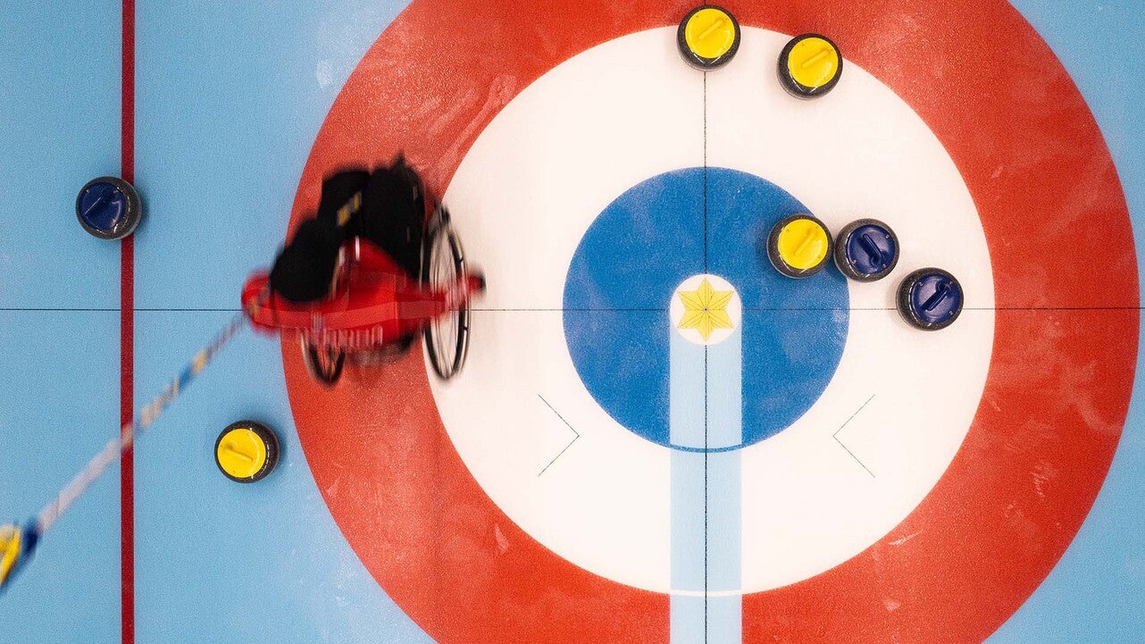 Fågelperspektiv över en curlingbana där en idrottare i rullstol skjuter en sten mot boet. Flera gula och blå curlingstenar ligger spridda på isen i målcirkeln.